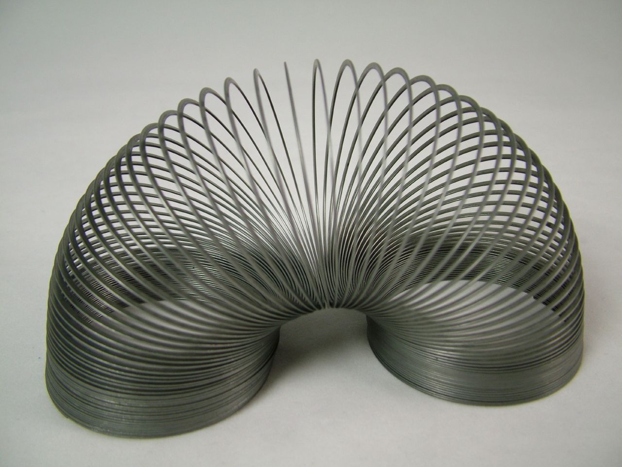 A black-and-white photo of a metallic helical spring (slinky) arranged in a curved shape on a reflective surface. The spring’s coils loop symmetrically outward from the center, forming a tunnel-like pattern with tightly wound turns, illustrating the spring bent into an arching shape.